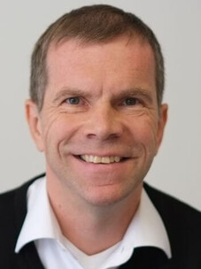 Porträt Friedrich Reinert reundlich lächelnd mit kurzem Haar und weißem Hemd vor neutralem Hintergrund.